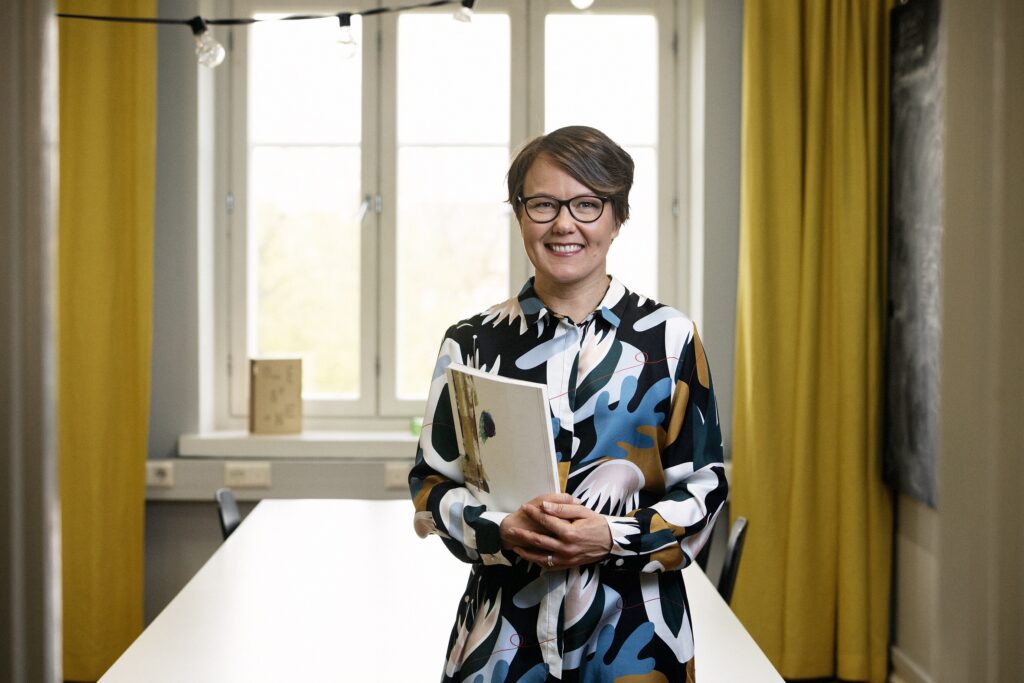 Kuvssa Heidi Korva seisoo huoneessa, ja taustalla näkyy palaveripöytä ja ikkuna, jossa on sinapinkeltaiset verhot